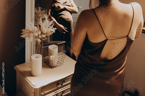Pregnant girl in a brown silk dress stands in the bedroom near the mirror and looks at the reflection. Mother in anticipation of the birth of a child, hugs her belly