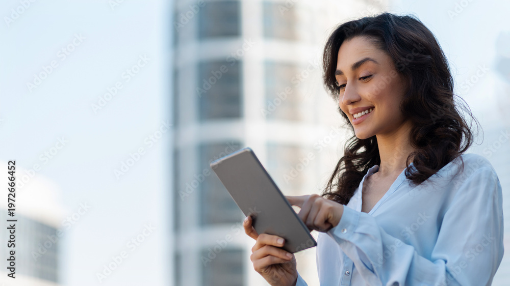 Fototapeta premium Panorama shot with businesswoman standing outside in urban area and using digital tablet while smiling and engaging with content, free space