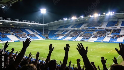 wide-angle photograph capturing the vibrant atmosphere of a large football stadium during a night match. The layout features a vast crowd of spectators in the foreground, with numerous hands raised in
