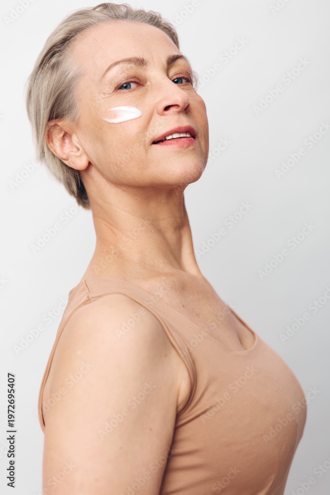 Fototapeta premium Mature woman with short grey hair applies white face cream on cheek. She wears beige top and poses confidently against a plain light background. Concept of skincare, aging, and self-care.