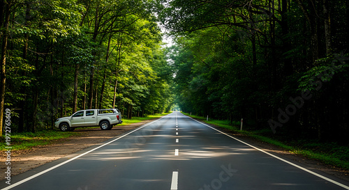 Straight asphalt road through lush green forest with white pickup truck parked. Scenic summer travel and road trip.