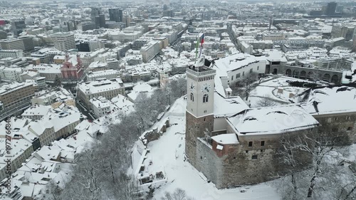 Lateral drone shot sweeps past the castle to uncover a wintry snow draped panorama of Ljubljana. Slovenia.