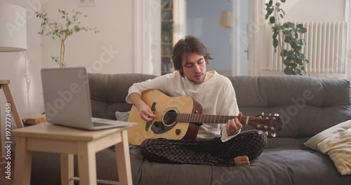 Strumming gentle tunes on his guitar while sitting comfortably on a sofa, filling the room with warm melodies