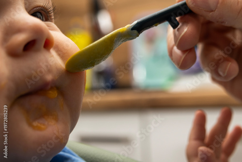 Фотография Close up of baby being fed with spoon, soft puree near mouth, capturing early feeding moment and gentle parenting care in warm natural indoor lighting