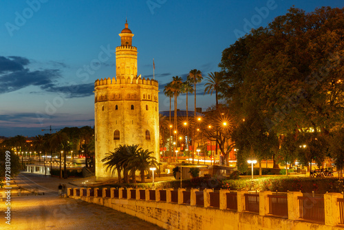 Evening view of Torre del Oro illuminated at dusk with palm trees and city lights in Seville, Spain.