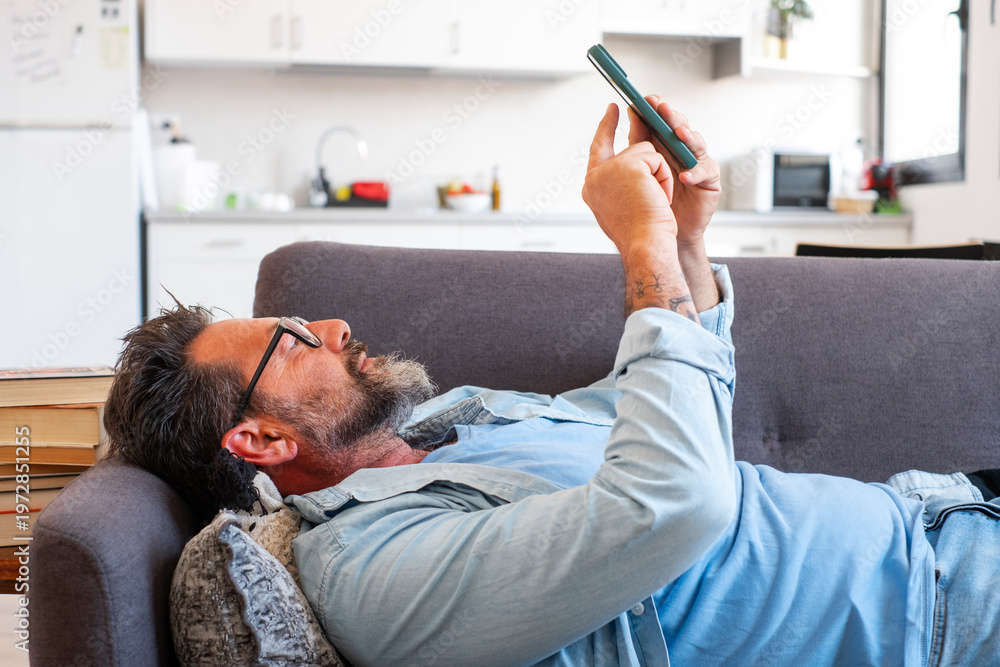 Fototapeta premium Man lying on sofa using smartphone with serious expression, focused and bored. Indoor lifestyle scene showing device dependence, scrolling, notifications and solitary digital consumption at home