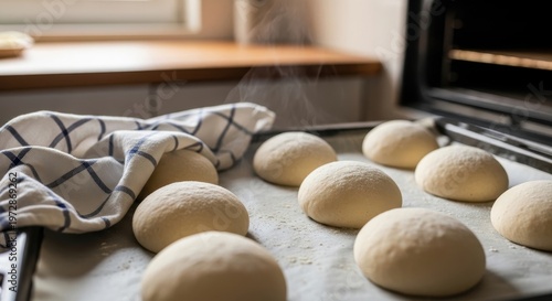 Freshly baked bread rolls on a baking sheet, ready to be put into the oven, with a kitchen towel nearby.