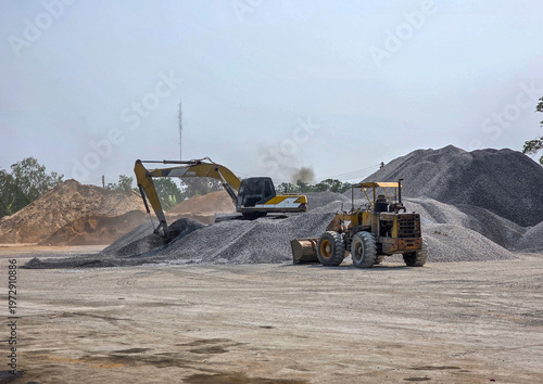 A hydraulic excavator and a wheel loader working together on large stockpiles of crushed stone at an industrial construction site.


