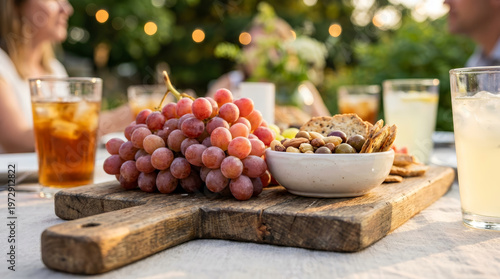 Grape appetizer charcuterie board outdoor picnic drink bunch of red grape beside snack bowl with nut and cracker rustic wooden board, outdoor picnic table with cold drink and blurred couple sunset