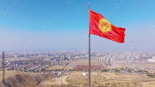 Waving Kyrgyzstani Flag Over Bishkek Cityscape - aerial view with truck to the right camera movement. Capital city buildings, homes, dry hills, hazy distance.