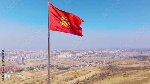 National Flag of Kyrgyzstan in Clear Weather in front of cityscape - wide orbiting drone view. Bishkek urban, suburbs, arid hills, distant haze.