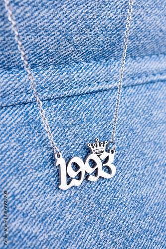 Close-up of a silver pendant featuring the year 1993 and a cute crown on a chain, set against a  blue  background.