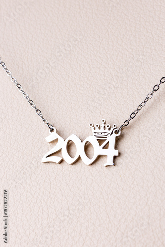 Close-up of a silver pendant with the year 2004 and a cute crown on a chain, set against a   luxurious white   leather background.