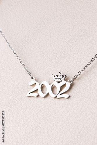Close-up of a silver pendant with the year 2002 and a cute crown on a chain, set against a luxurious white leather background.