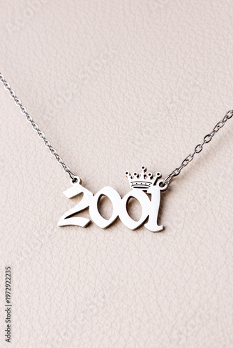 Close-up of a silver pendant with the year 2001 and a cute crown on a chain, set against a luxurious white leather background.