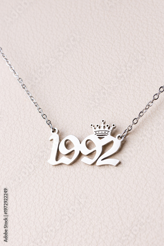 Close-up of a silver pendant with the year 1992 and a cute crown on a chain, set against a luxurious white leather background.