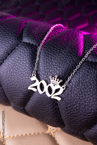 Close-up of a silver pendant with the year 2002 and a cute crown on a chain, set against a luxurious black leather background.