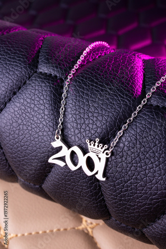 Close-up of a silver pendant with the year 2001 and a cute crown on a chain, set against a luxurious black leather background.