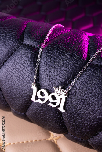 Close-up of a silver pendant with the year 1991 and a cute crown on a chain, set against a luxurious black leather background.