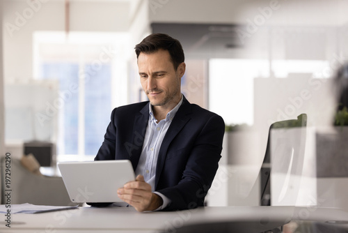 Canvas Print Focused adult Hispanic businessman reviewing digital and paper documents reading online report browsing web for information comparing data planning project using tablet pc at office desk
