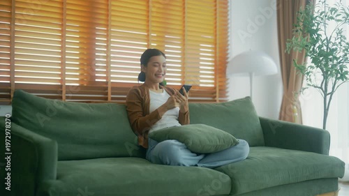 Happy young asian woman relax on comfortable couch at home texting messaging on smartphone, smiling girl use cellphone, chatting online message, shopping online from home
