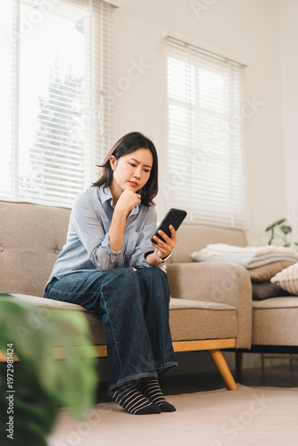 Concerned Asian woman sitting on a sofa and looking at her smartphone in a bright living room