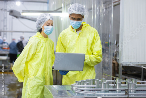 Billede på lærred Two factory workers in protective uniforms and face masks inspect canned food production line with laptop in manufacturing
