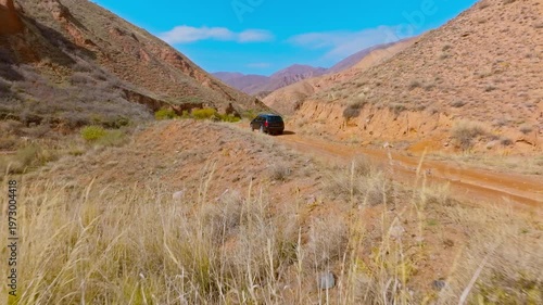 Vehicle navigates a rugged dirt road through eroded sandstone mountains in Kyrgyzstan. Daytime adventure captures the essence of remote journeys. Drone view.