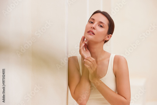 woman gently touching her neck with fingertips, looking upward with a serene expression, leaning against a white column. Editorial beauty portrait, copy space left. I