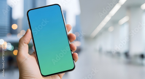 Close-up of a hand holding a modern smartphone with a blank blue-green screen in a blurred office or building interior background.