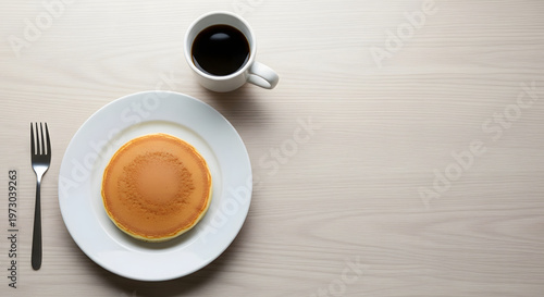 A single golden pancake on a white plate with a fork and a cup of black coffee on a light wooden table, viewed from above.