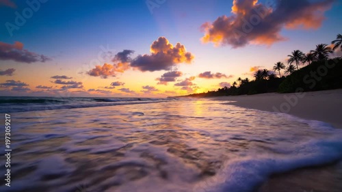 A tropical beach at sunset or sunrise with orange clouds reflecting on wet sand, gentle ocean waves approaching the shore, and silhouetted palm trees on a coastal hill under a blue sky.
