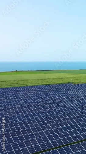 Wide solar panel installation in a green field overlooking the sea under a clear blue sky. Renewable energy concept in a natural coastal landscape.