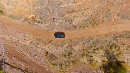 Dark-colored vehicle travels along a rugged dirt road in an arid desert landscape. Directly above drone view.