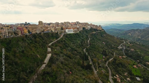 Aerial view of Enna, Sicily showcasing the scenic hillside cityscape with winding roads, lush greenery, and historical architecture under a cloudy sky