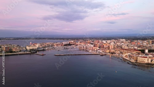 Aerial view of Ortigia at dawn, showcasing the tranquil harbor, colorful buildings, and soft pastel sky reflecting on the calm water surface