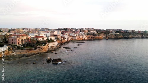 Aerial view of the coastal city of Syracuse, Italy, showcasing colorful buildings along the shoreline with calm waters and rocky formations in the foreground