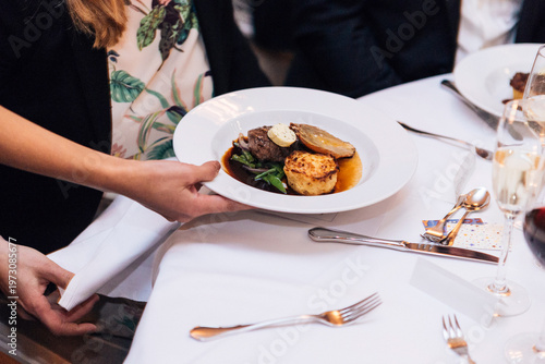 Elegant waiter serving a gourmet beef steak dinner with potato gratin, roasted onion, and red wine sauce at a formal banquet. High-end catering, fine dining restaurant service, and luxury events.