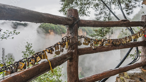Many different padlocks are mounted on a chain on the wooden railing of the observation deck. Behind the fence, you can see mountains, shrouded in fog and clouds, trees. China. Zhangjiajie. Avatar 
 