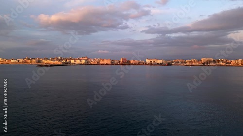 Ortigia at dawn, showcasing the gradual illumination of buildings along the waterfront, with soft reflections on the calm water and a pastel sky enhancing the serene atmosphere