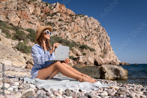 Woman, laptop, beach. Young woman remotely working on a sunny pebble beach with rocky mountains and copy space.