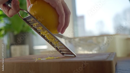 Close up of hands grating fresh lemon zest with metal grater on wooden cutting board in kitchen. Concept of cooking baking preparation and homemade food.