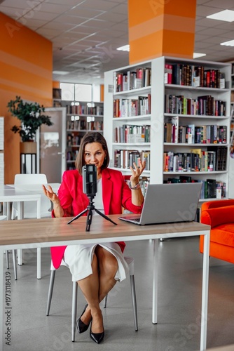 smiling professional woman recording video on smartphone with tripod and laptop in library interior looking at camera content creator online education business communication, vertical