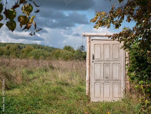 Freestanding weathered wooden door stands alone at the edge of an open field. The surreal scene evokes themes of escape, transition, and unexpected possibilities