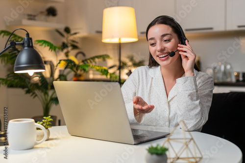 Young woman at night home works as call center agent with headset answers support call and logs ticket. Caucasian girl on sofa at evening listens speak note request polite tone service aid quick log.