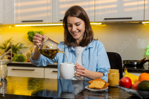 Cuadro en lienzo Young woman pours herbal tea into mug in home kitchen indoors feeling relaxed after meal prep