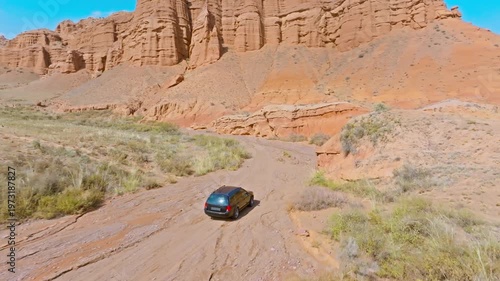 Off-road vehicle navigates rugged dirt road through arid desert canyon. Vast landscape evokes feelings of adventure and exploration. Following drone view.