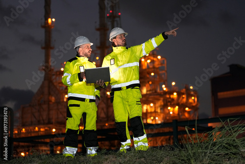 Engineers in high-visibility gear inspect industrial plant at night with laptop