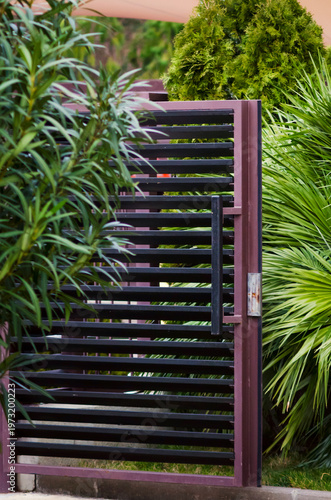 A modern brown metal entrance gate surrounded by decorative bushes. Vertical photo.