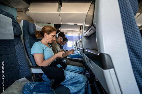 Quadro em tela Passengers are sitting in economy class airplane seats, using their personal sma